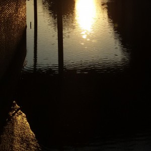 Fotografie eines Fleets, in dessen kräuselndem Wasser sich die untergehende Sonne spiegelt.