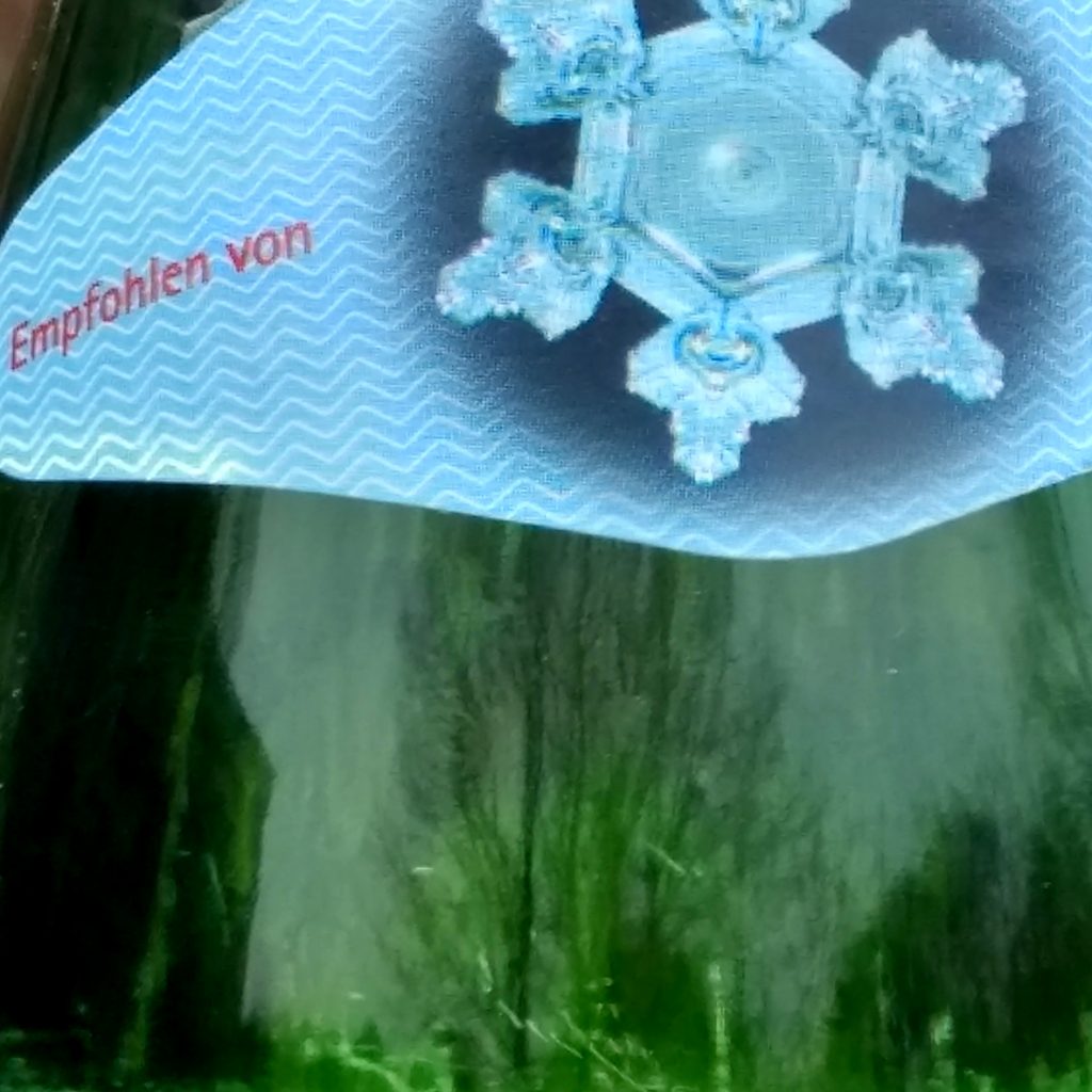 Ein Frühlingsgarten spiegelt  sich im grünen Glas einer Wasserflasch, auf deren Etikett eine Schneeflocke prangt.